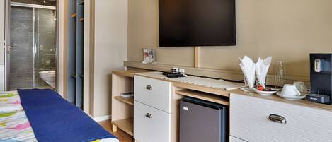 A modern hotel room with a shower, a bed with a blue blanket, a desk with a coffee machine, and a flat-screen TV.