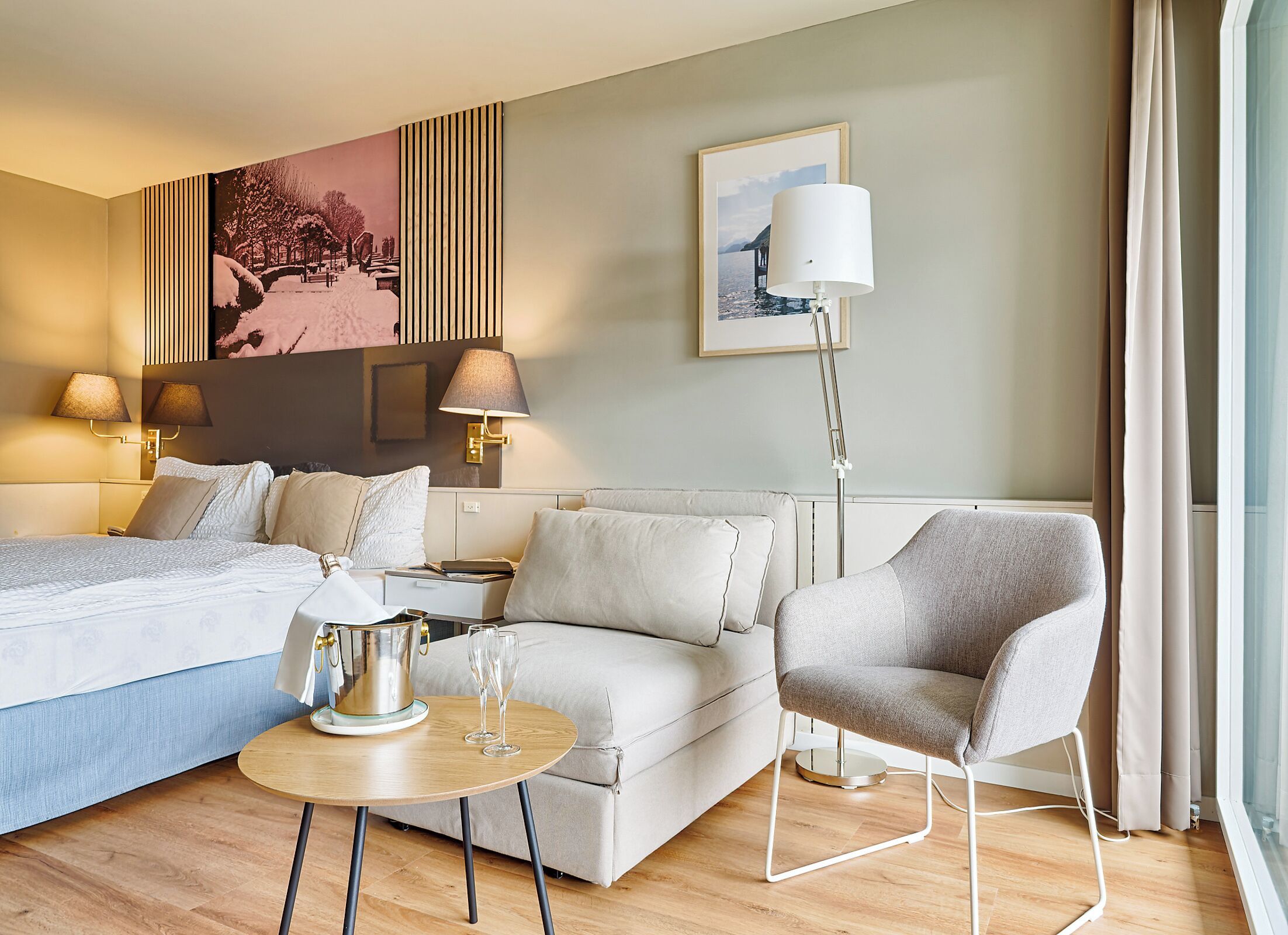 A hotel room with a bed, a sofa, a chair, a table with glasses, a lamp, and artwork on the walls.