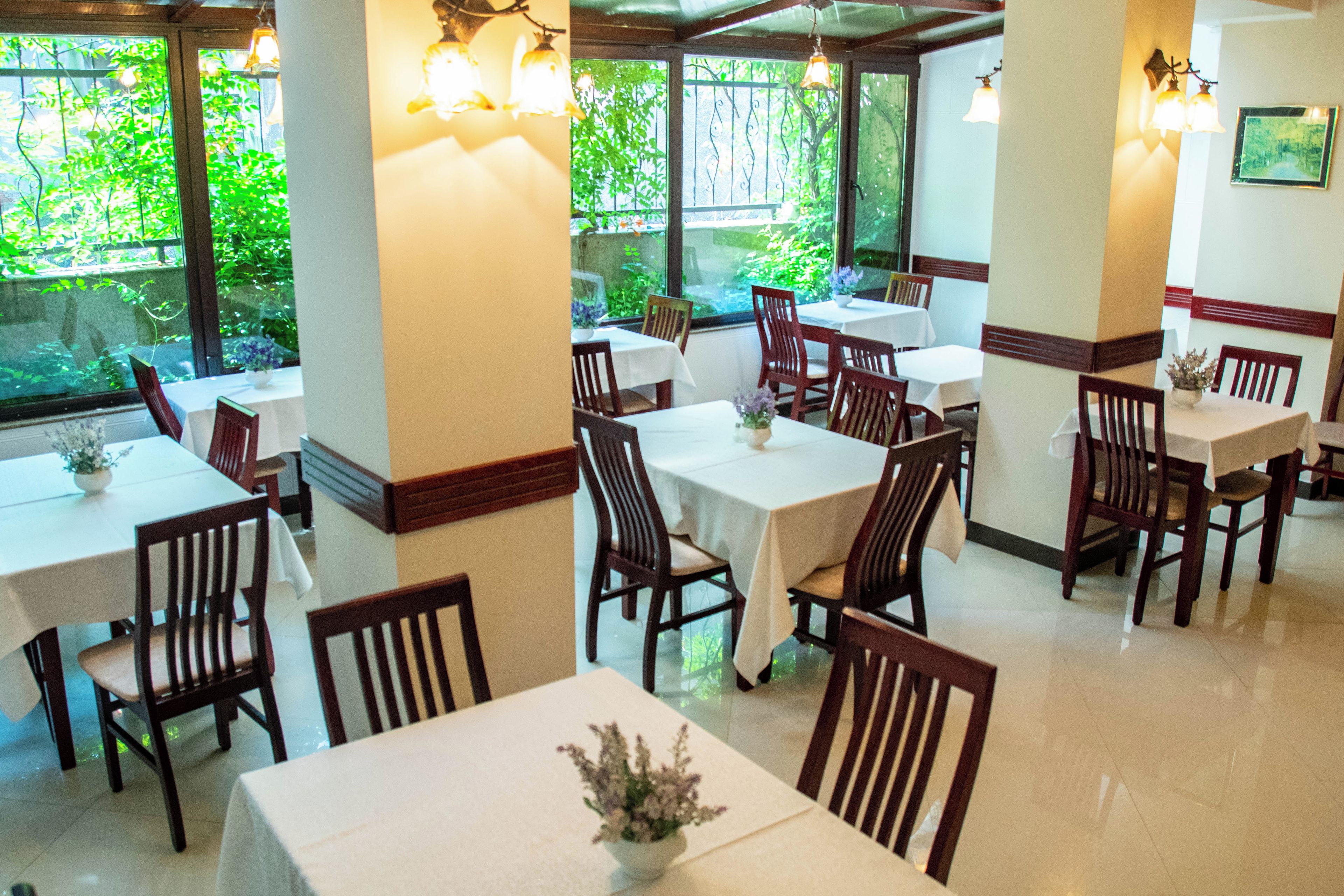 A restaurant interior with multiple tables and chairs, large windows with greenery outside, and wall-mounted lighting.