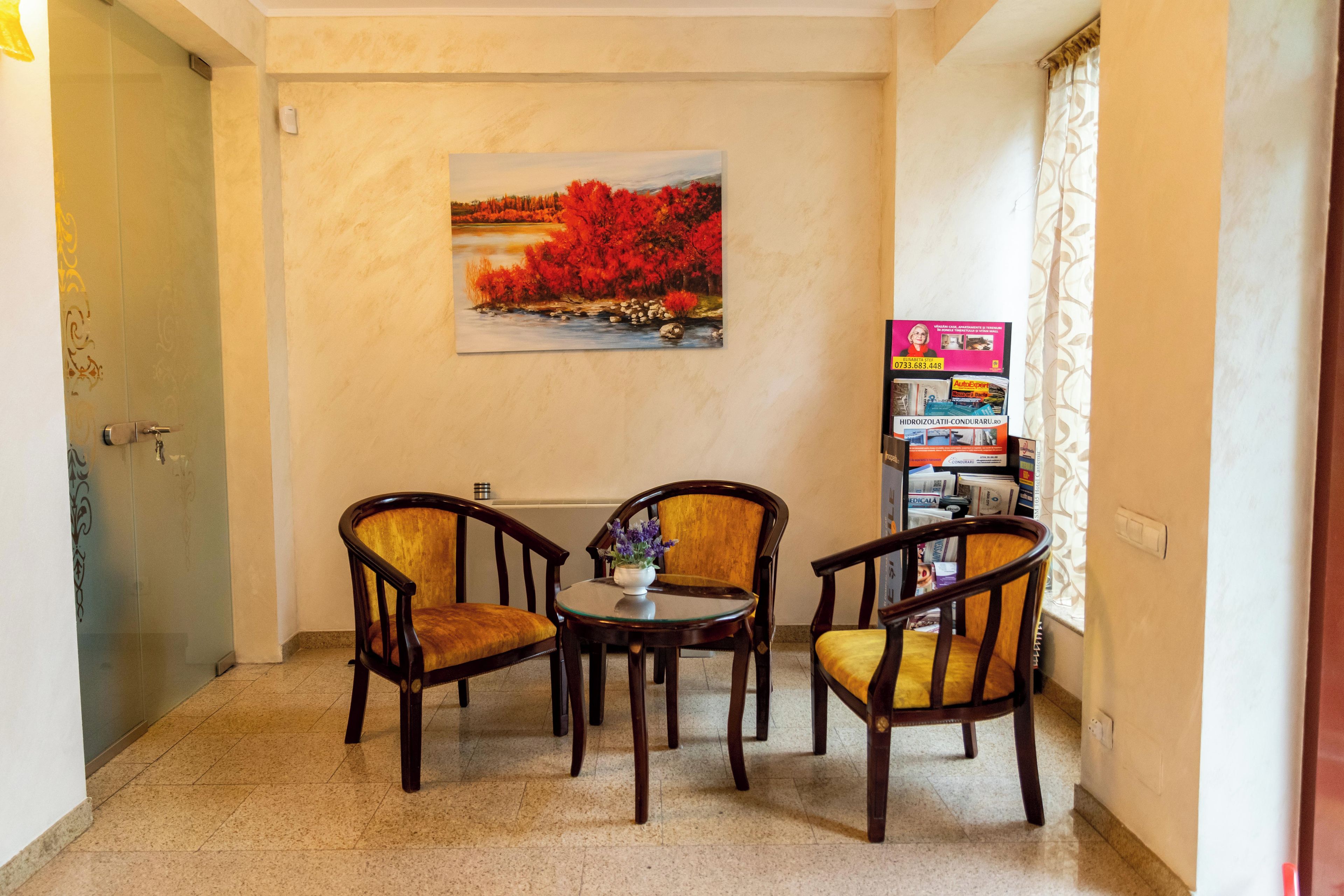 Quarto com pintura de paisagem, duas cadeiras de madeira, uma mesa pequena e uma estante com revistas.