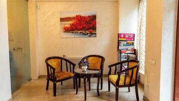 Quarto com pintura de paisagem, duas cadeiras de madeira, uma mesa pequena e uma estante com revistas.