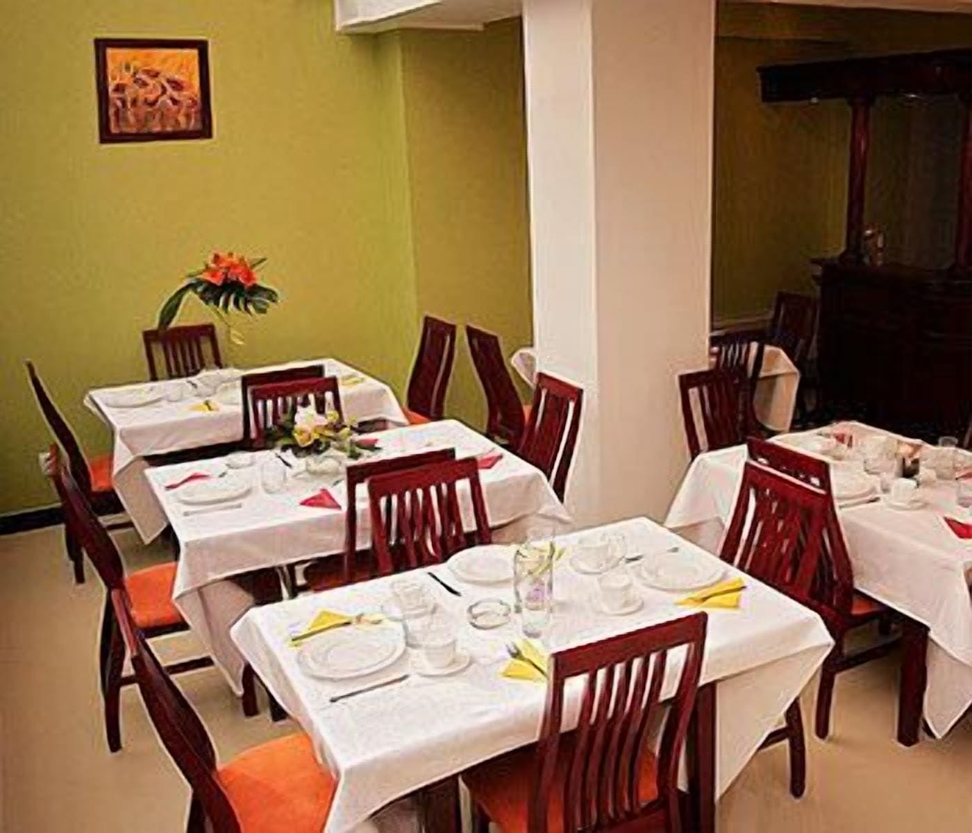 A dining area with tables set for four, each with white tablecloths, red chairs, and a vase of flowers.