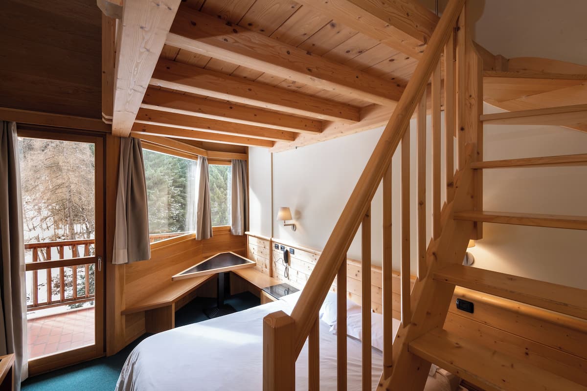 A wooden cabin room with a bed, a desk, and a window overlooking a balcony.