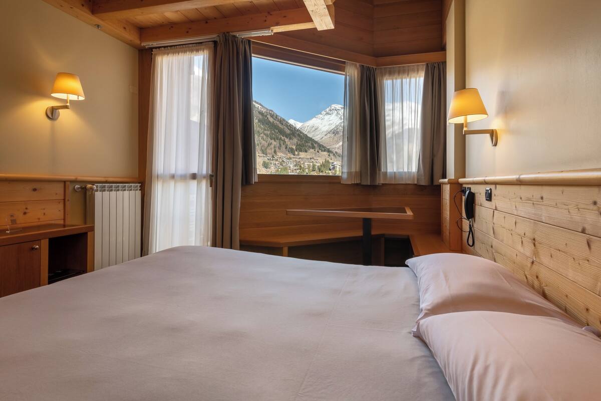A bedroom with a large bed, wooden headboard, two bedside lamps, a window with curtains, and a view of mountains.