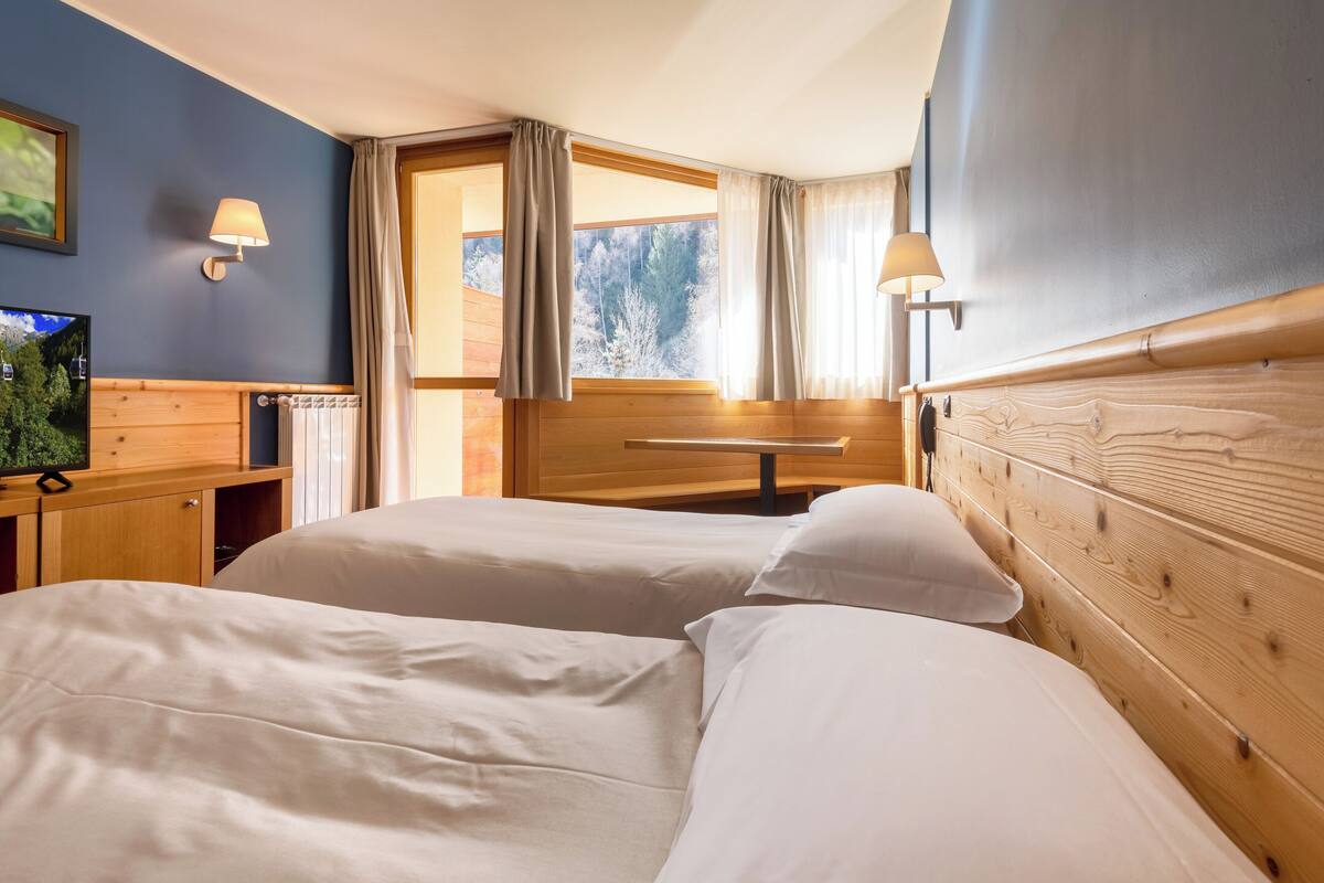 A hotel room with two beds, wooden headboard, a wooden desk with a TV, and a window with curtains.