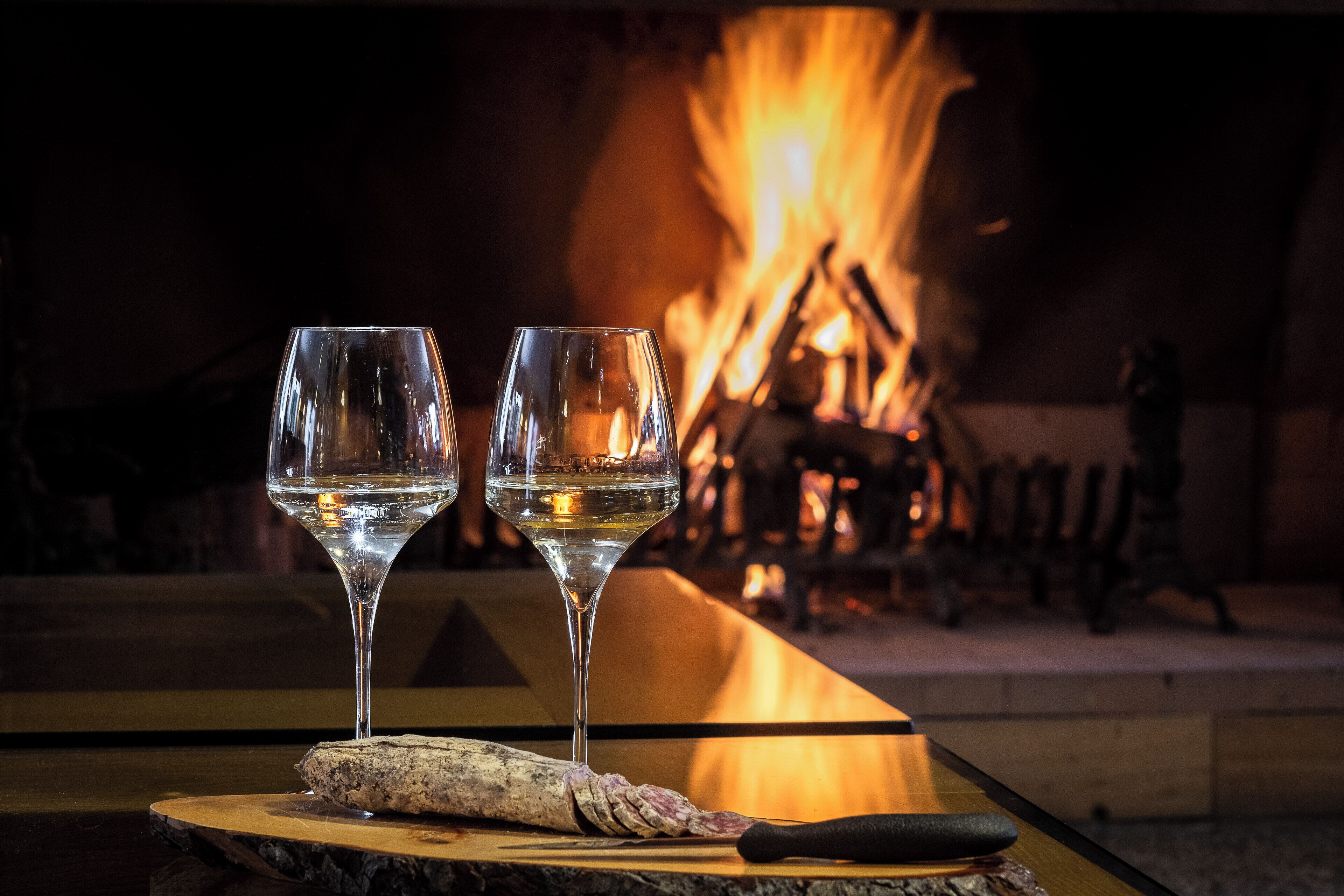 Two glasses of white wine on a wooden table with a log and a knife, in front of a fireplace.