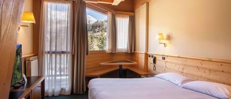 A hotel room with a wooden ceiling, a bed, a desk with a lamp, and a view of the mountains through the window.