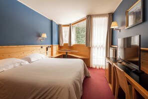 A hotel room with a bed, a desk with a TV, a lamp, and a window with curtains. - Hotel Garnì Pegrà (Ponte di Legno)