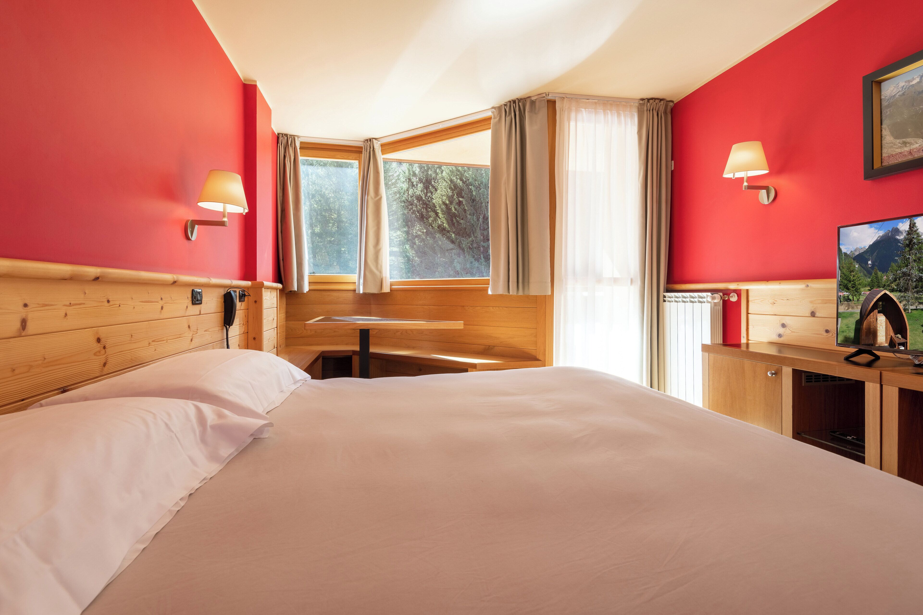 Quarto de hotel com uma cama grande, cabeceira de madeira, duas luminárias de apoio, uma escrivaninha de madeira, televisão e uma janela com cortinas.