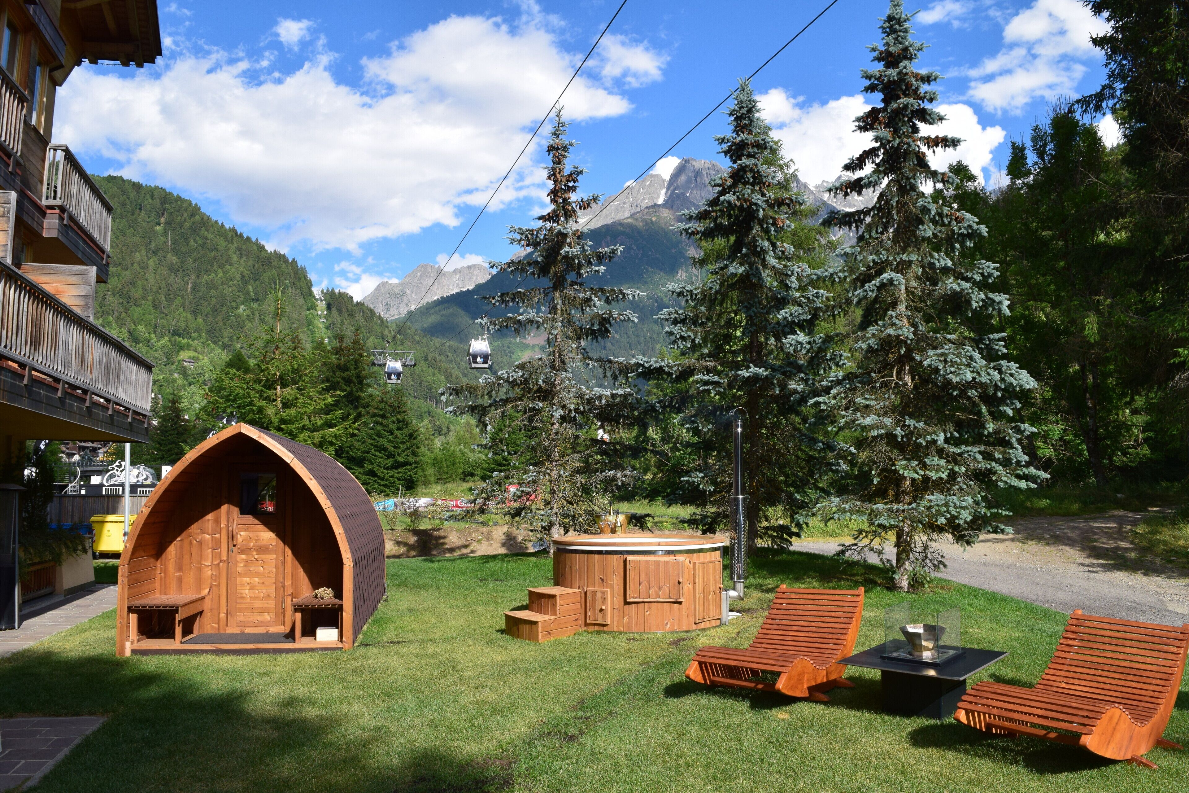 Eine Holzhütte mit geneigtem Dach, einem Whirlpool und zwei Liegestühlen auf einer Wiese, im Hintergrund Berge.