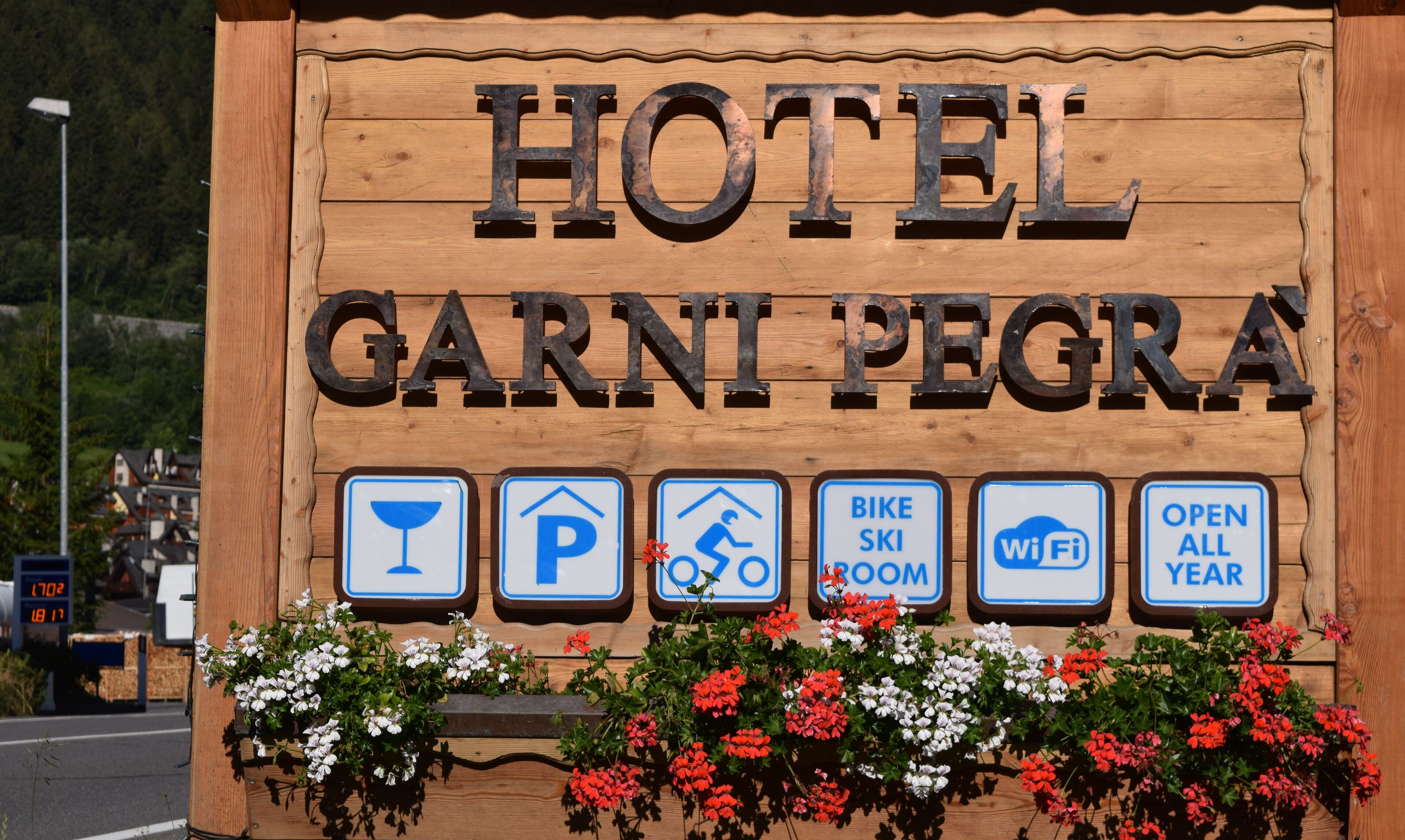 Une enseigne en bois indiquant l’hôtel Garni Pegria, avec des pictogrammes représentant les équipements disponibles : parking, local à vélos, local à skis, connexion Wi-Fi et ouverture toute l’année.