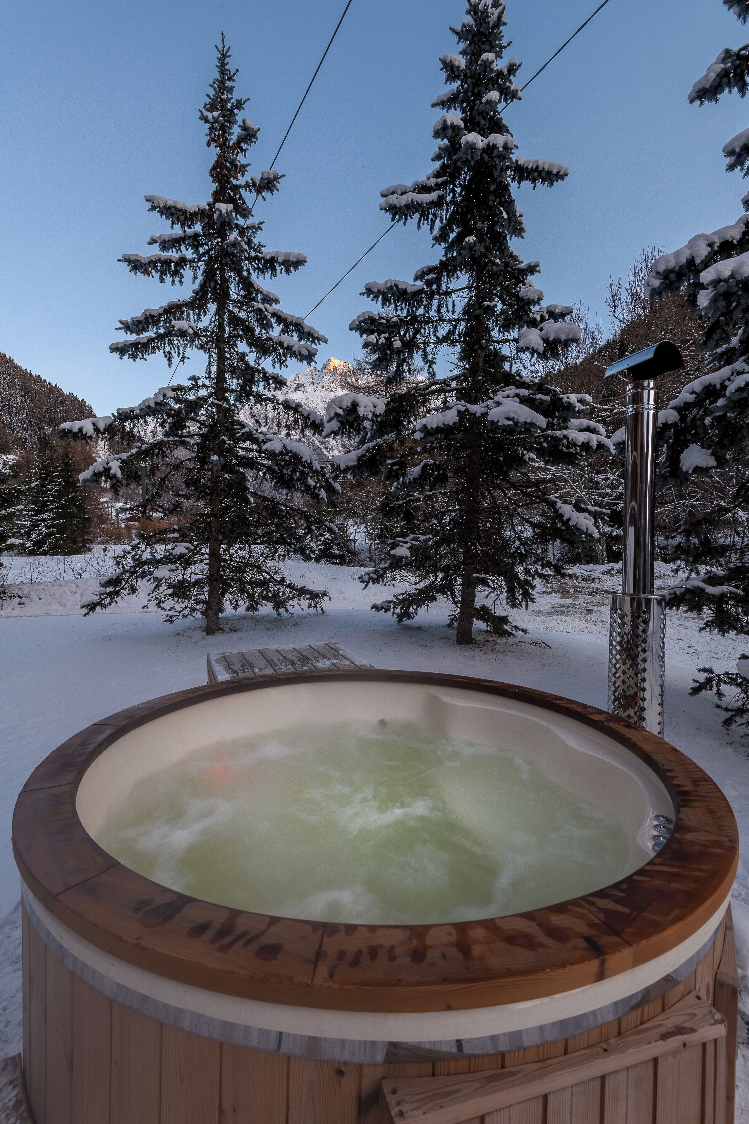 A wooden hot tub surrounded by snow-covered trees and mountains.