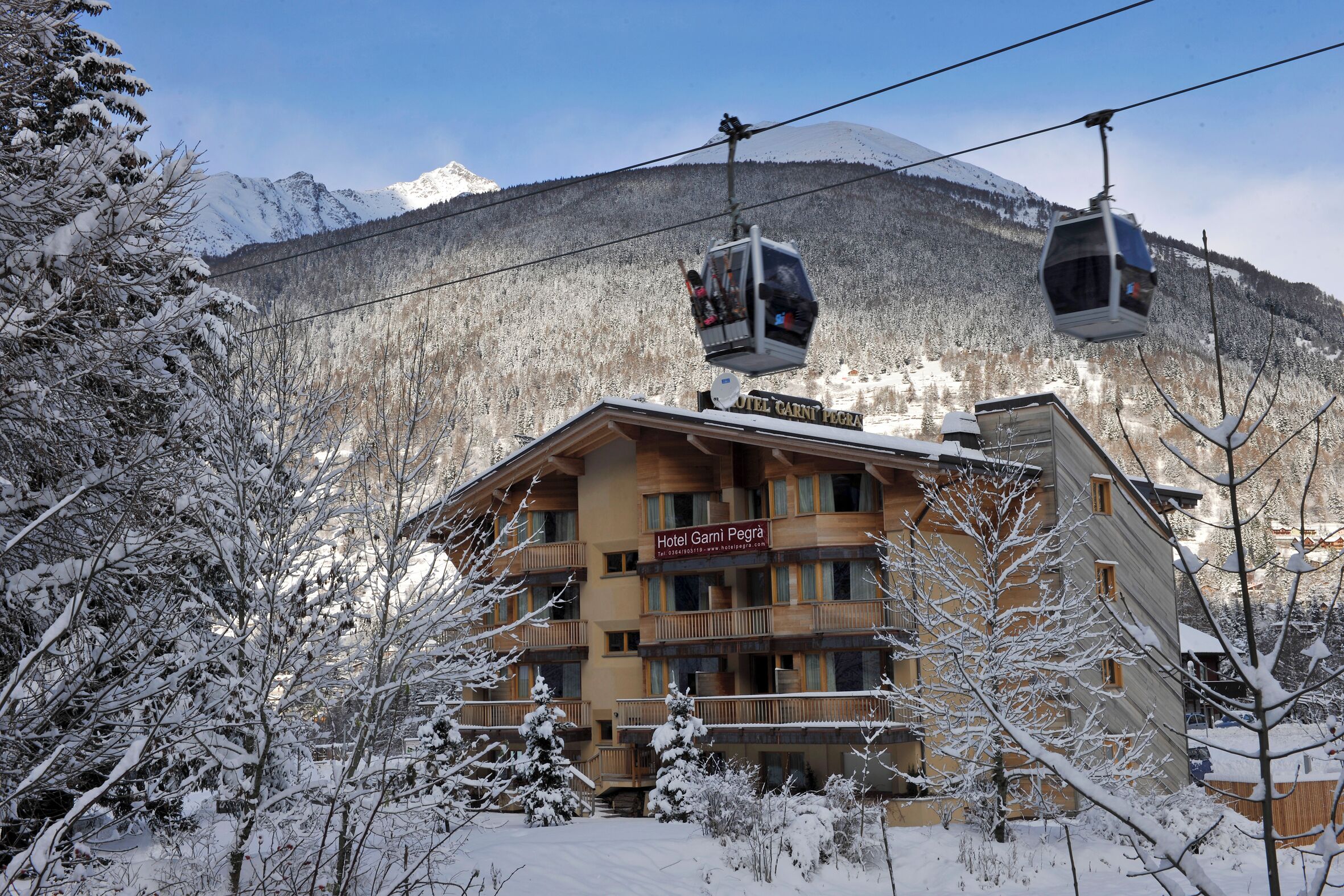 Ein Hotel mit einem Schild, auf dem 'Hotel Garni Pegria' steht, in einer verschneiten Berglandschaft mit einer Seilbahn im Hintergrund.