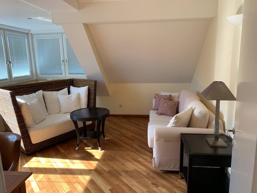 A spacious loft apartment with a high ceiling, a large window with blinds, a brown wicker sofa, a white sofa, a small round table, and a lamp on a side table.