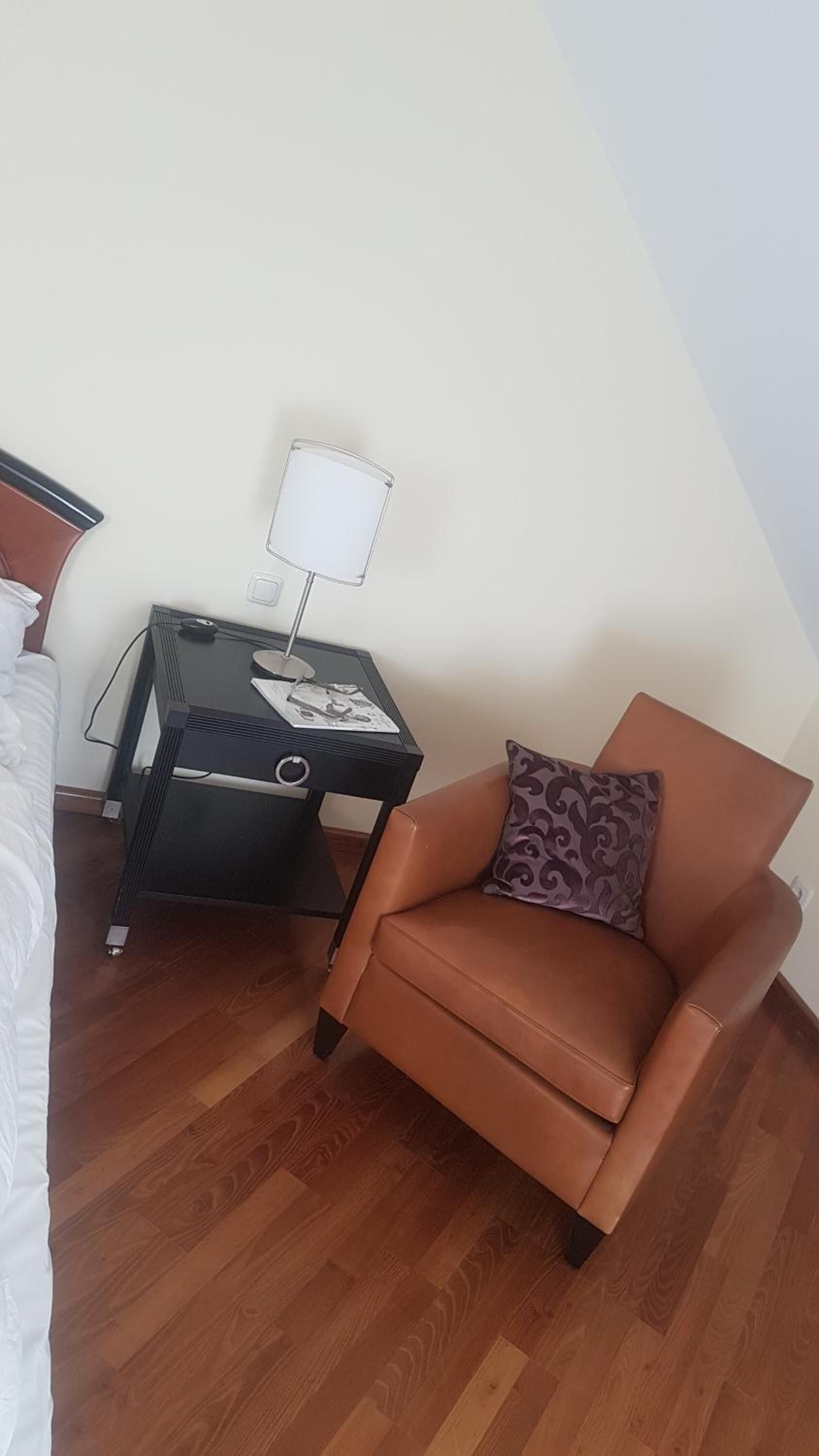 A hotel room with a brown leather armchair, a small black side table with a lamp, and a white bed.