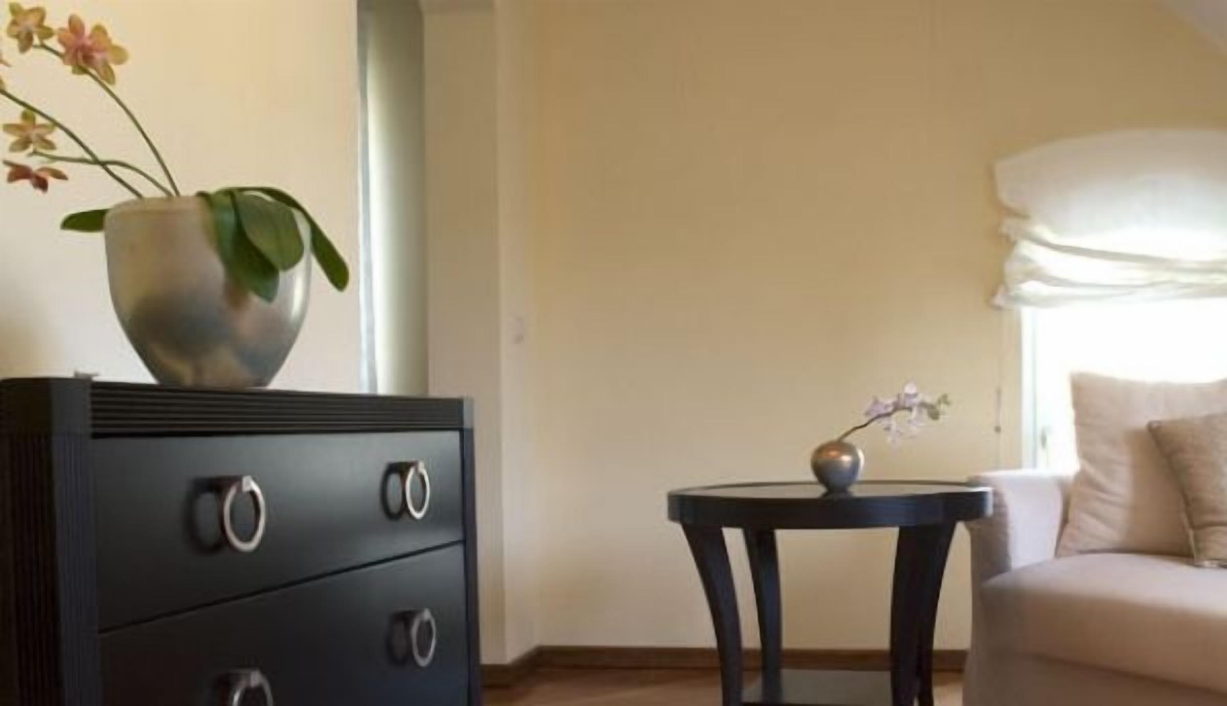 Una stanza con una cassettiera scura con tre cassetti, un tavolino rotondo con un vaso di fiori e un divano dai toni chiari.