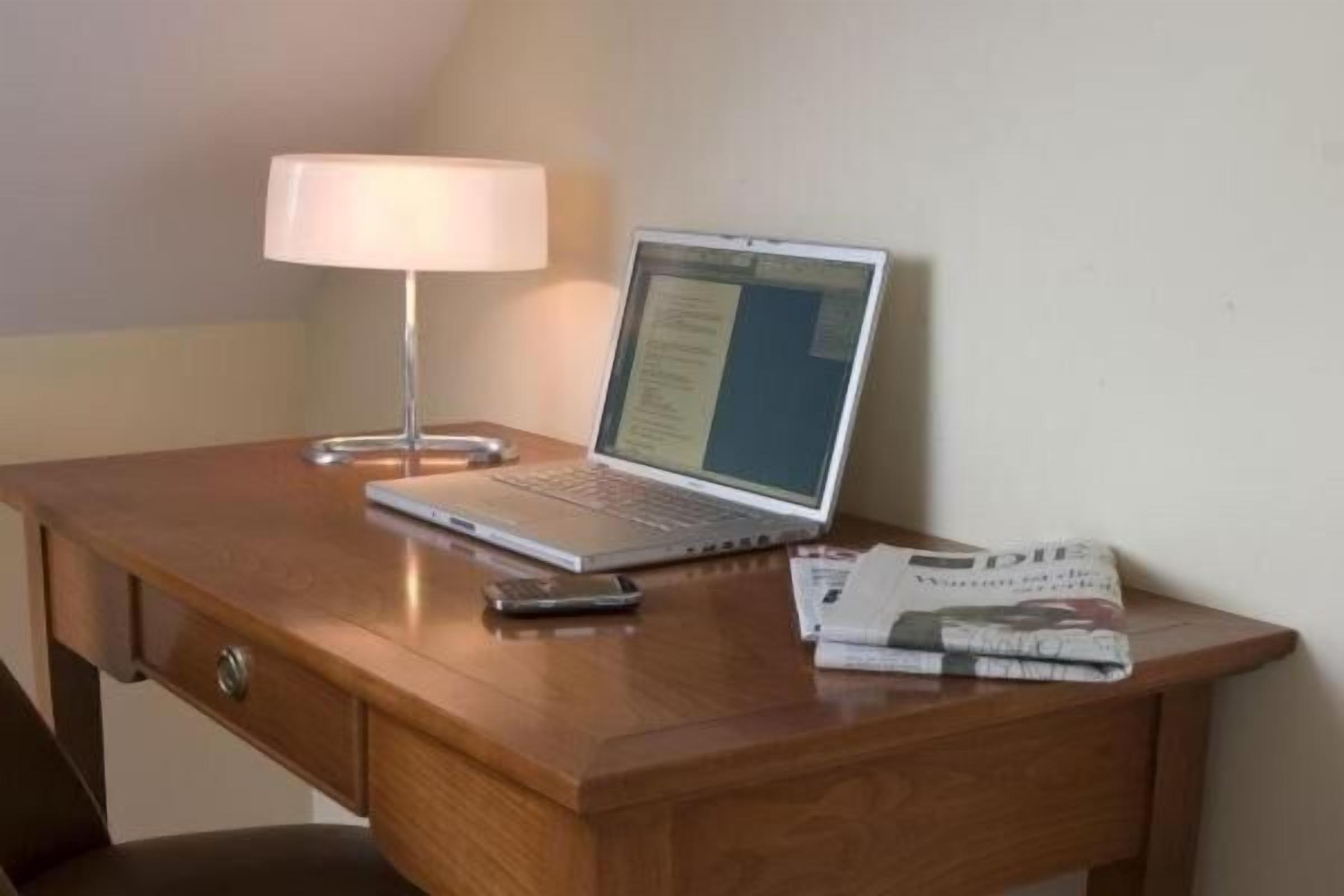 A wooden desk with a laptop, a lamp, a smartphone, and a magazine.