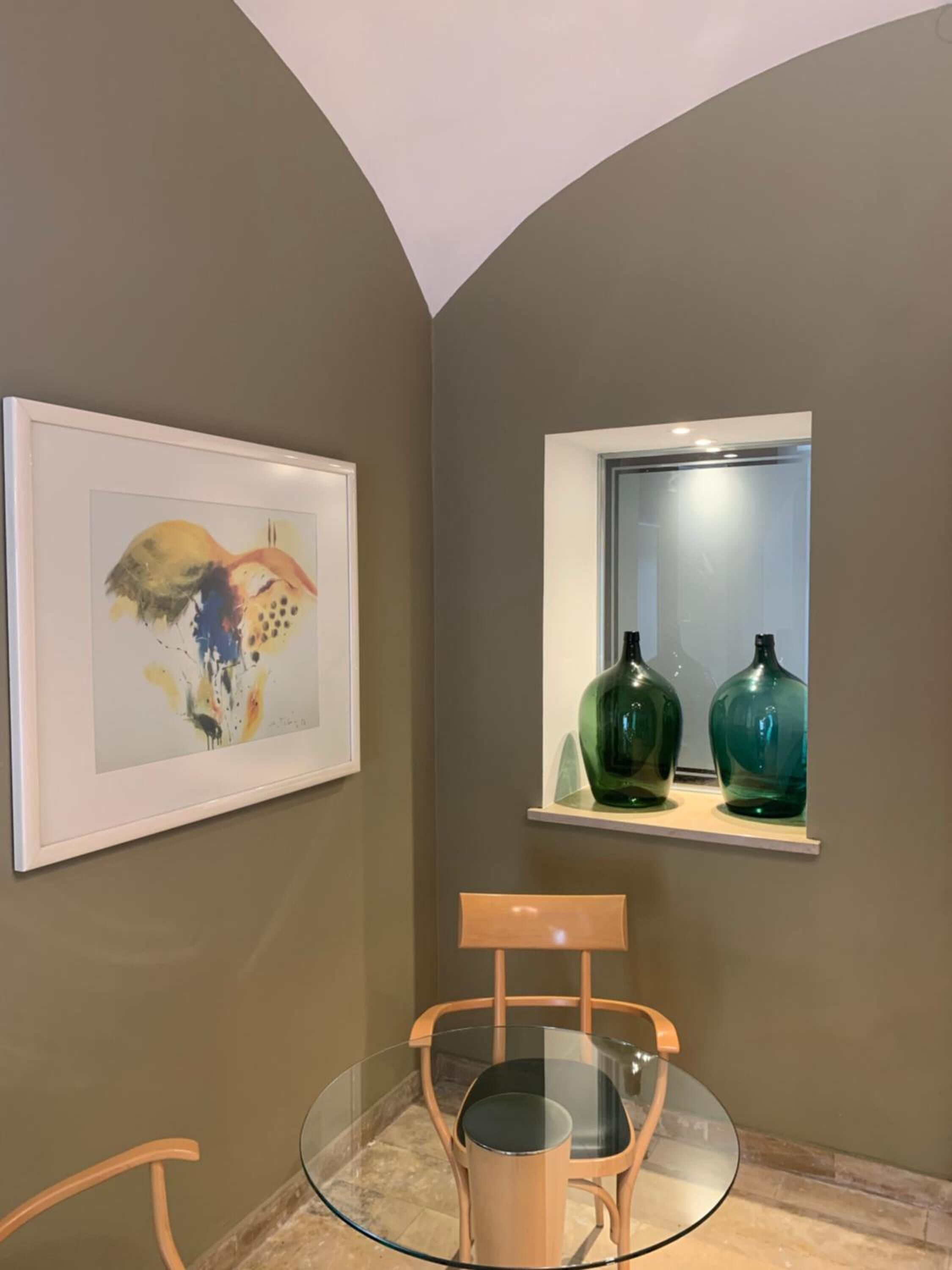 A room with a glass table and chairs, a framed abstract painting, and two green vases on a shelf by a window.