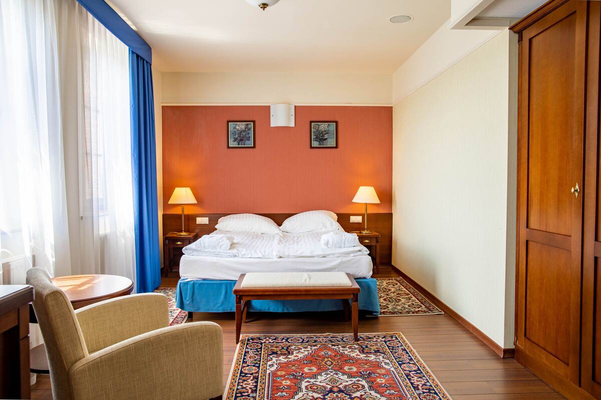 A hotel room with a bed, two bedside lamps, a wooden bench, a small table, and a patterned rug.