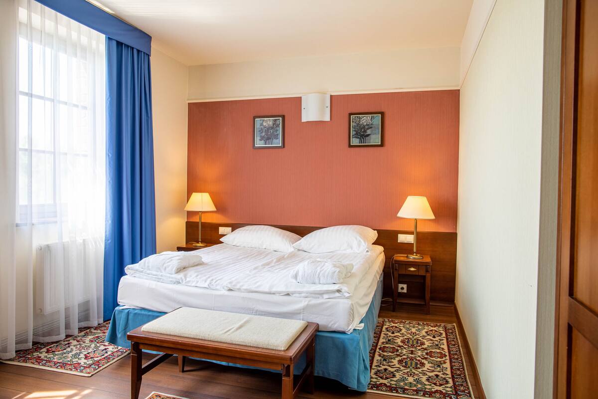 A hotel room with a bed, two bedside lamps, a bench, a patterned rug, and a window with blue curtains.
