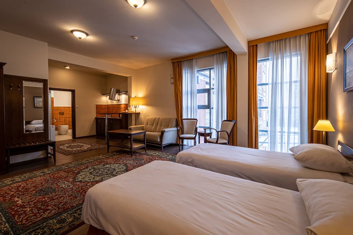 A hotel room with two beds, a desk, a chair, a sofa, and a large window with curtains.