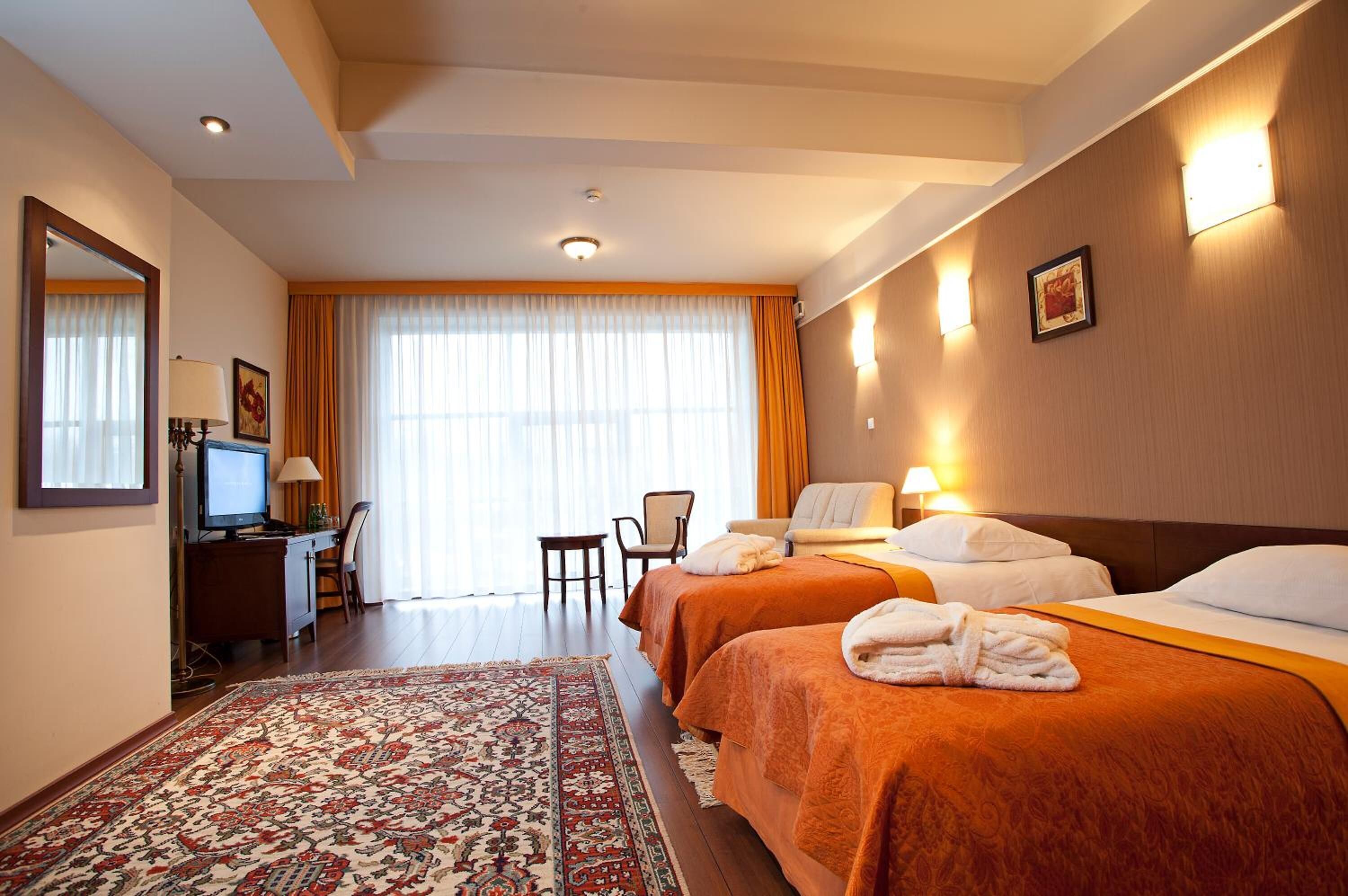 A hotel room with two beds, a desk with a computer, a chair, a large window with curtains, and a patterned rug.