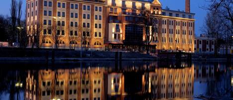 Un gran hotel de varios pisos y una chimenea destacada, reflejado en aguas tranquilas.