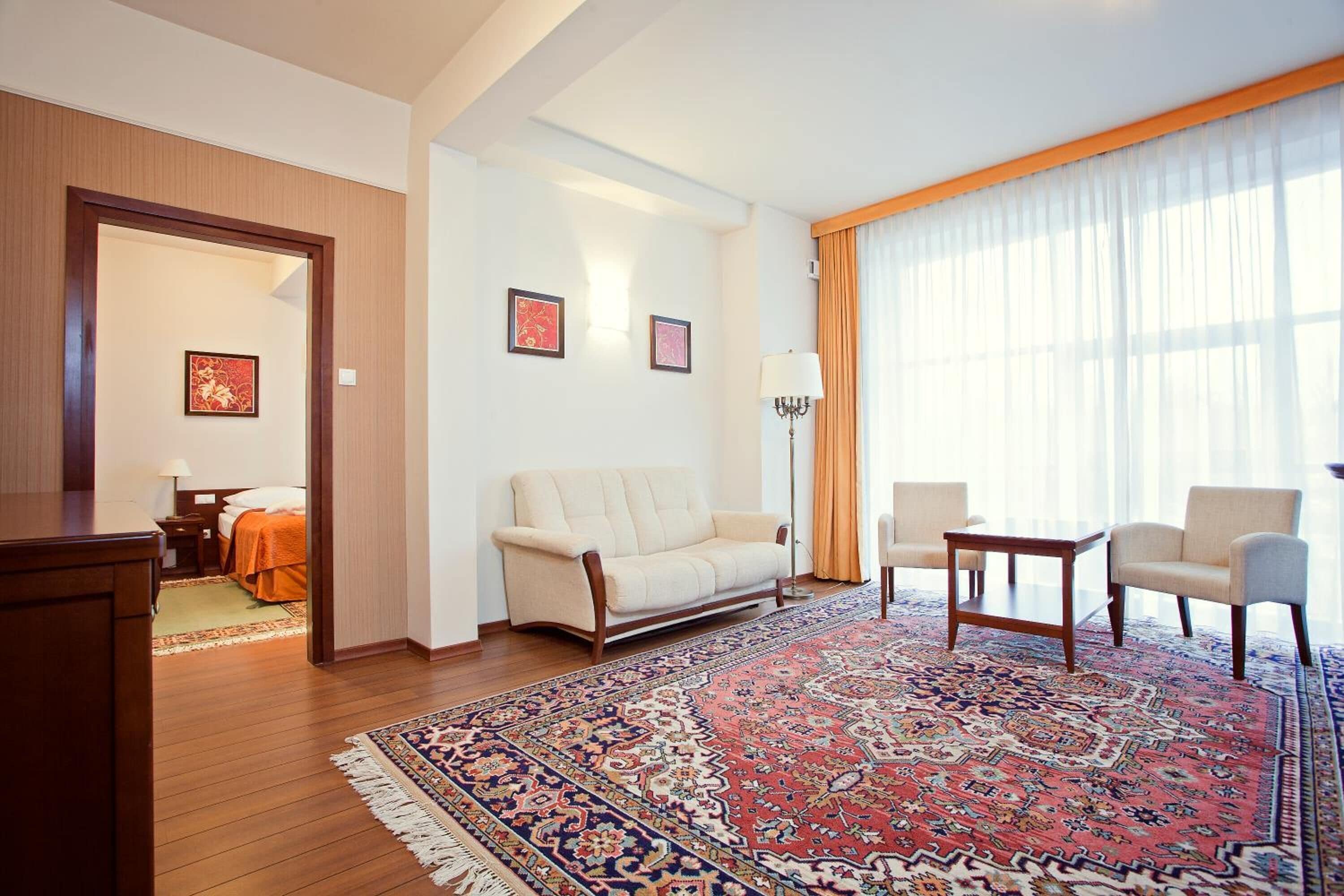 A hotel room with a large window, a sofa, two armchairs, a coffee table, a rug, and artwork on the walls.