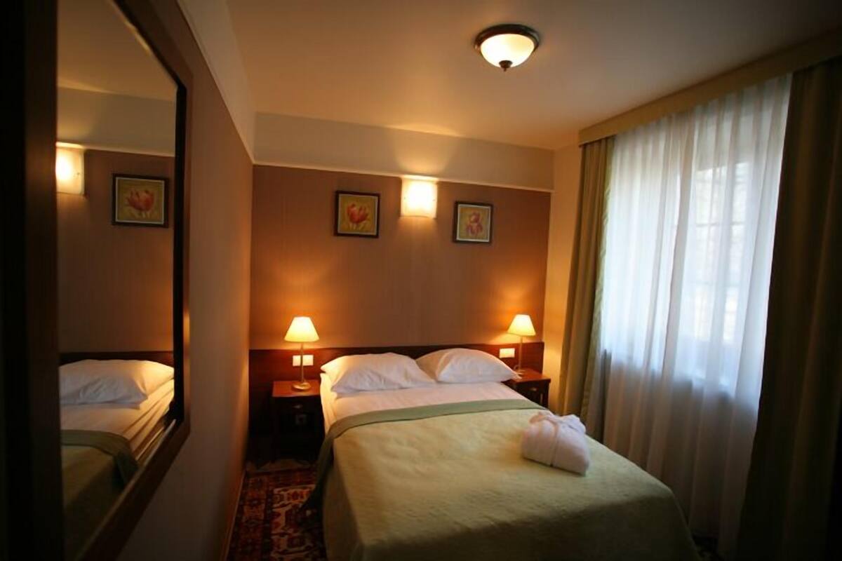 A hotel room with a single bed, two bedside lamps, a mirror, and a window with curtains.