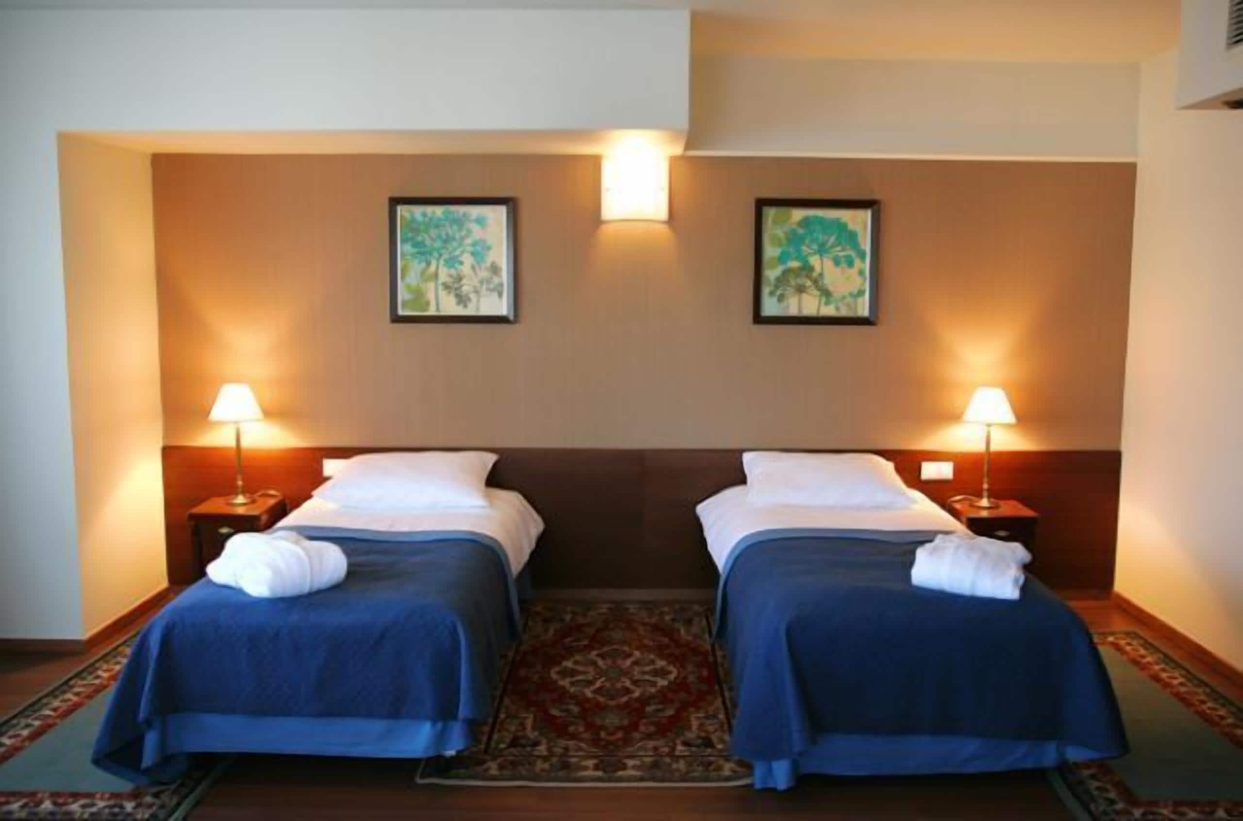 A hotel room with two beds, a large rug, and two wall-mounted lamps.