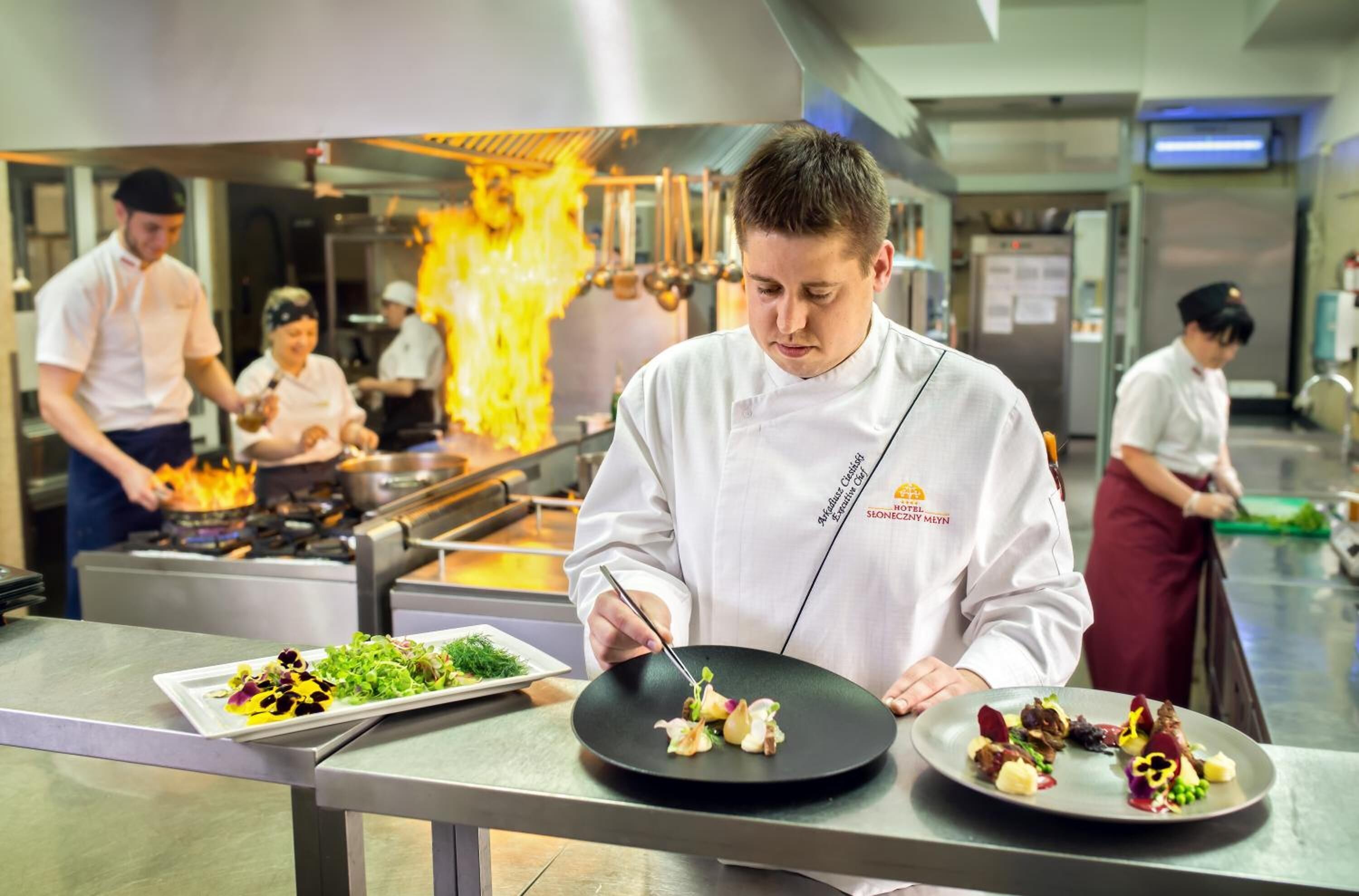 A chef preparing food in a professional kitchen with stainless steel surfaces and a visible fire in the background.