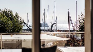 Un puerto deportivo con varios veleros amarrados, un gran crucero al fondo y vista al agua a través de una ventana con una silla y una mesa en primer plano.