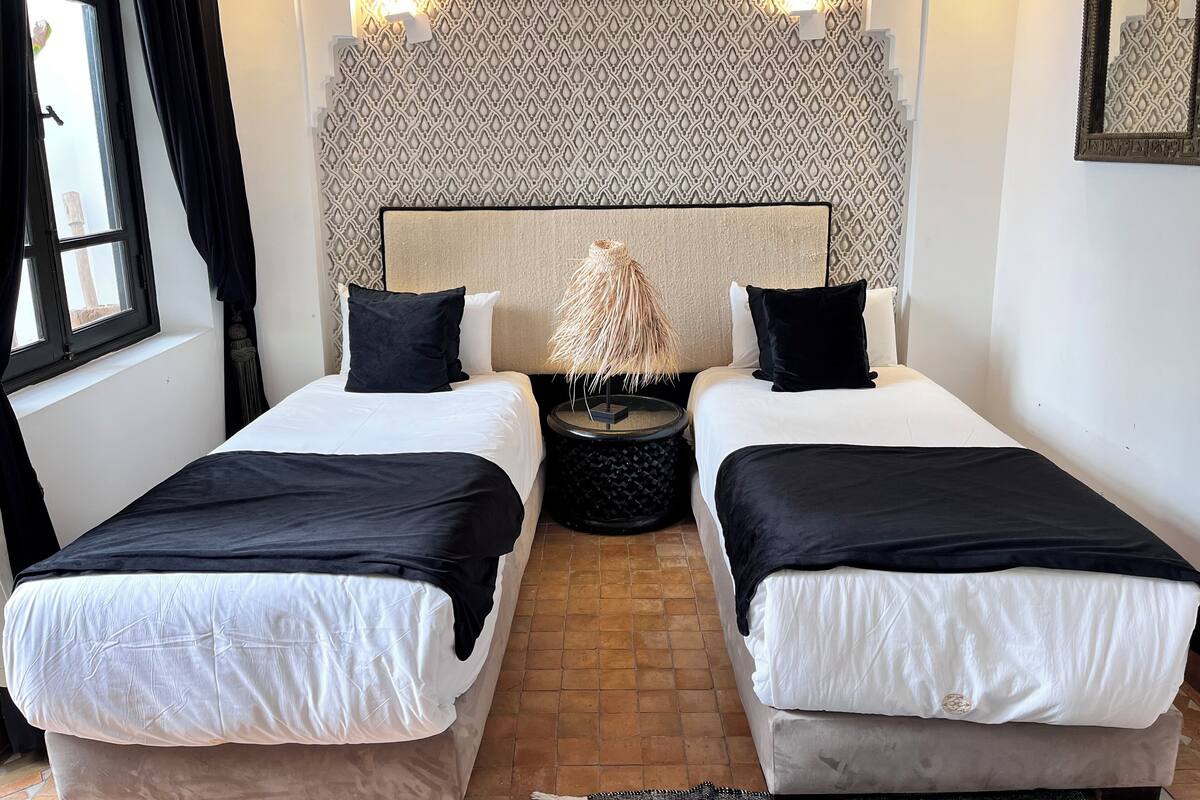 Habitación de hotel con dos camas individuales, un cabecero estampado, una mesita de noche con lámpara y una ventana con cortinas.
