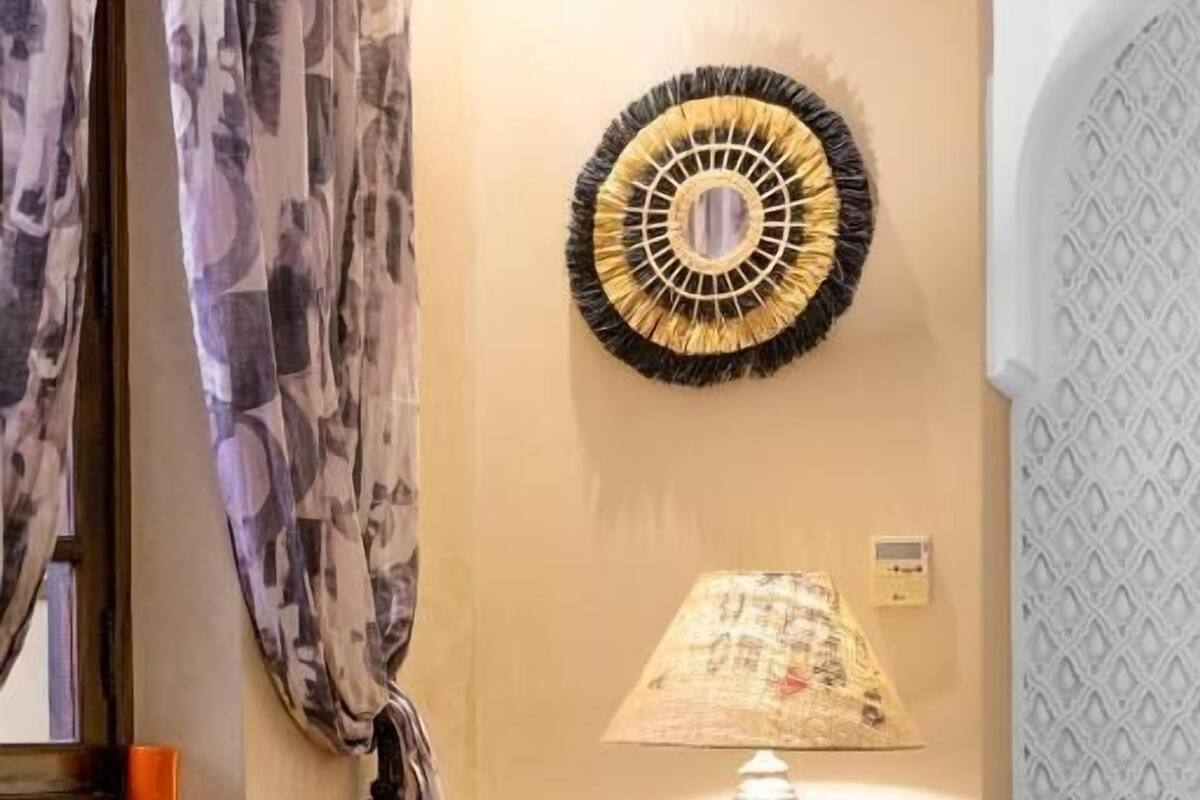 Una habitación de hotel con cama, una mesita de noche con lámpara, un adorno de pared y una cortina.