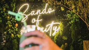 En hand håller i ett glas, och i bakgrunden syns en neonskylt med texten ”Sando of Unity”.
