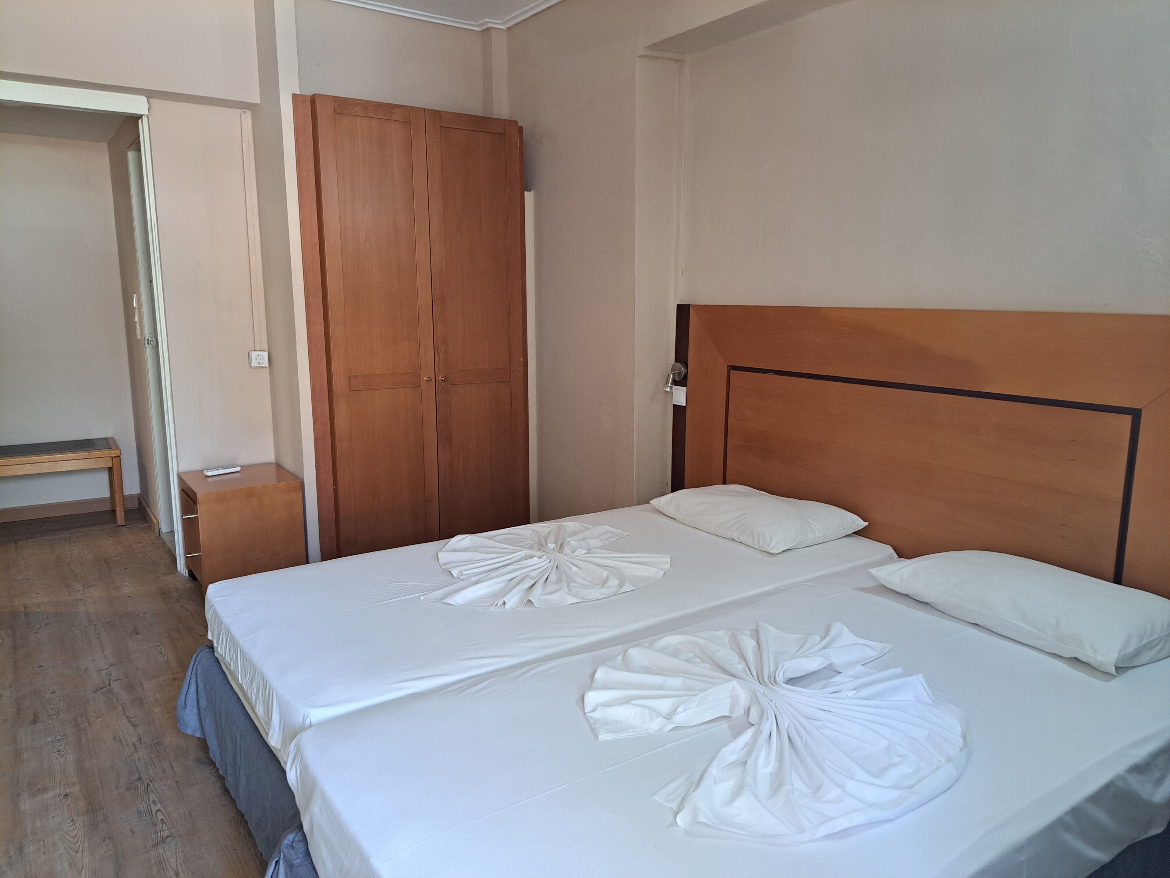 Una cama bien hecha con sábanas blancas y cabecera de madera, un armario de madera y una mesita de noche con un control remoto.