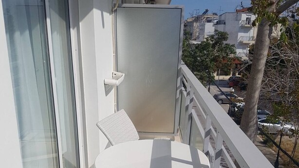 Un balcón con una mesa blanca y dos sillas de mimbre, con vistas a una calle con coches aparcados y edificios.