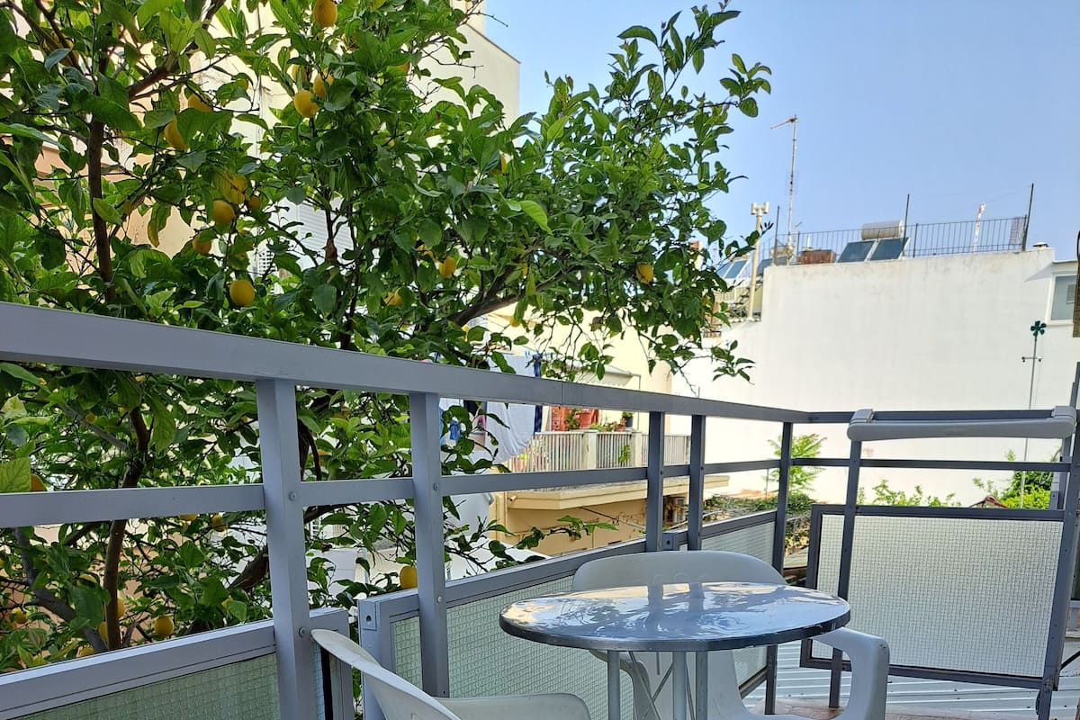 Terraza con una mesa, dos sillas, barandilla metálica y vistas a un edificio con paneles solares.