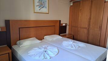 A hotel room with a wooden headboard, two beds with white linens, a nightstand, a wooden wardrobe, and a framed picture on the wall.
