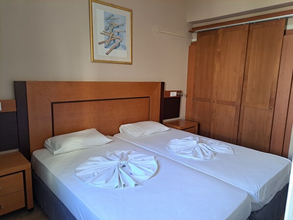 A hotel room with a wooden headboard, two beds with white linens, a nightstand, a wooden wardrobe, and a framed picture on the wall. - Bristol Hotel & Apartments (Kos)