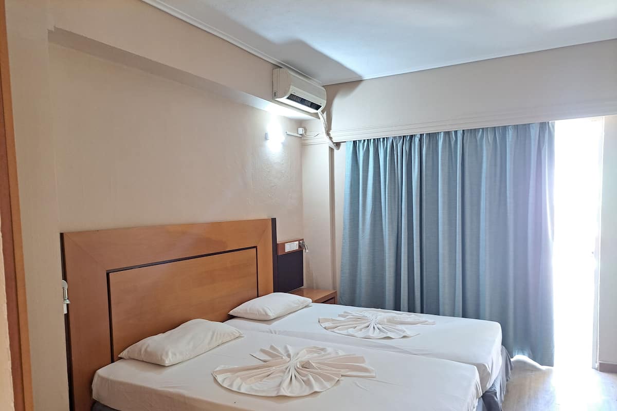 Habitación de hotel con dos camas, cabecero de madera, aire acondicionado en el techo, ventana con cortinas azules y piso de madera.