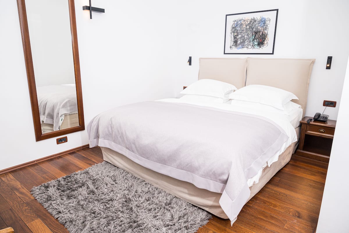 A neatly arranged bedroom with a large bed, a bedside table, a nightstand, a mirror, and a framed artwork on the wall.