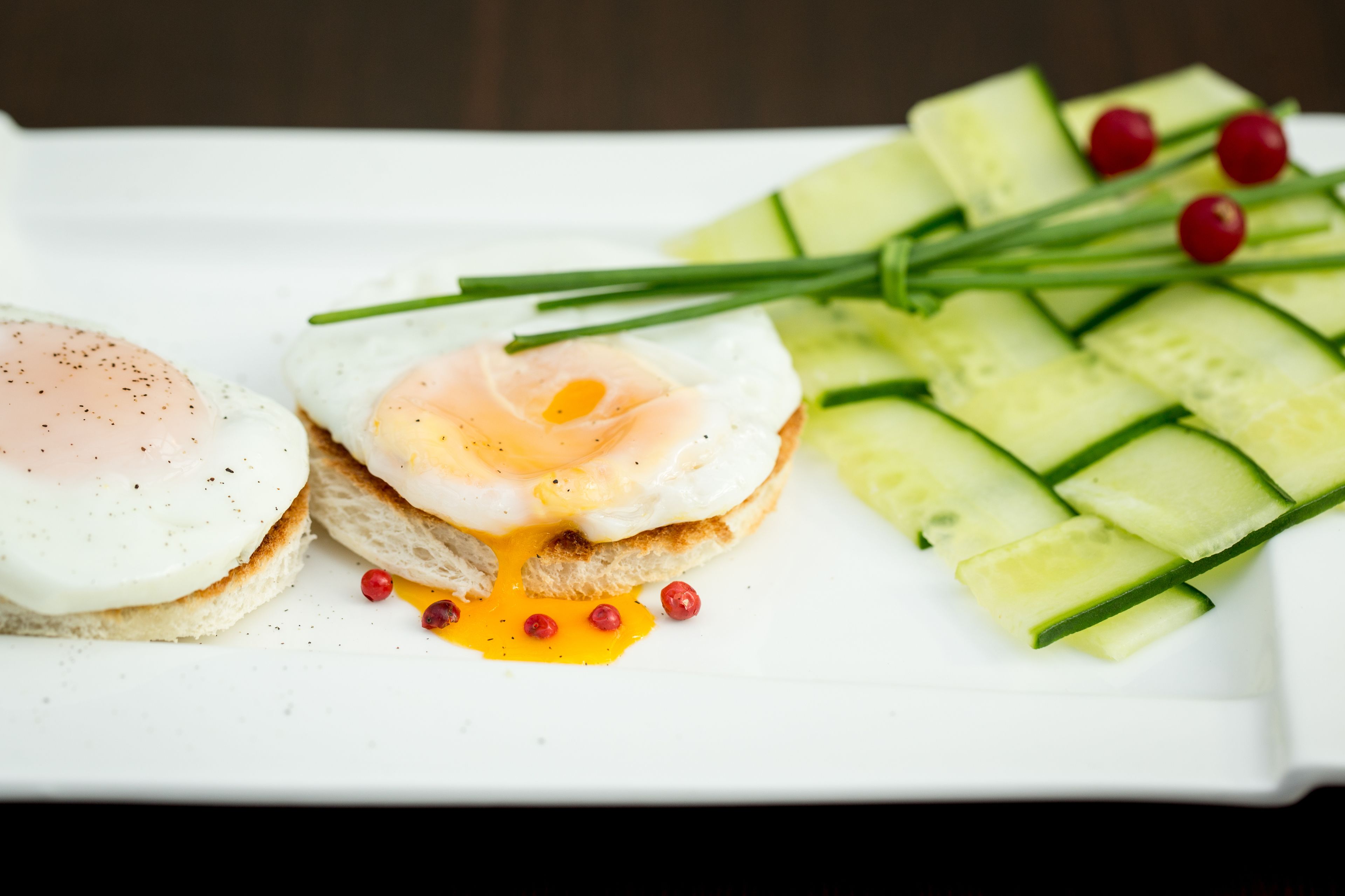 A breakfast plate with two poached eggs, a slice of toast, cucumber slices, chives, and red peppercorns.