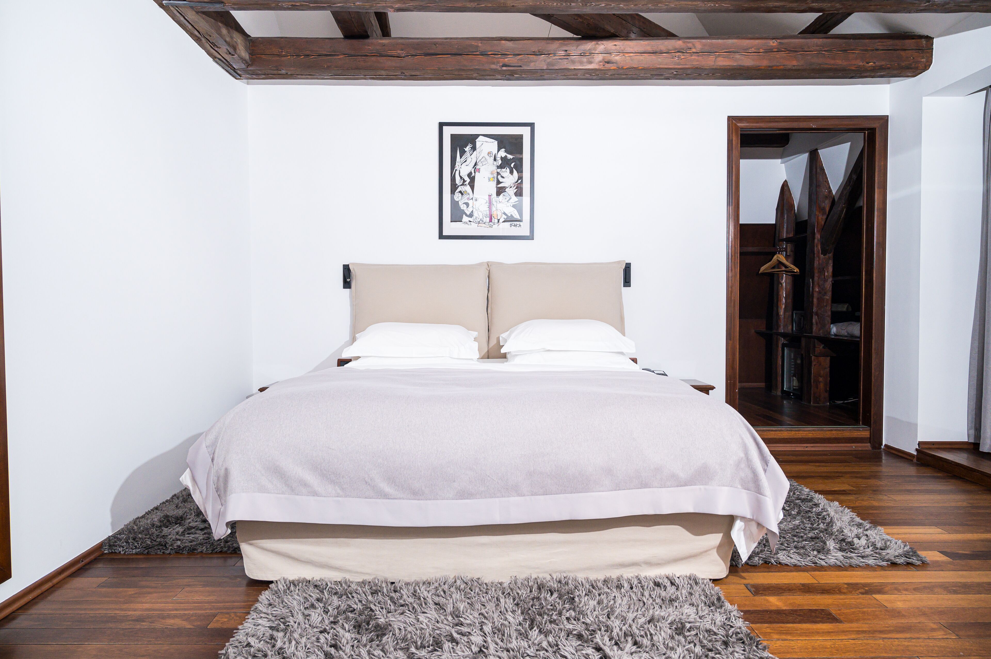A bedroom with a large bed, wooden beams on the ceiling, a framed picture on the wall, and a wooden door leading to another room.