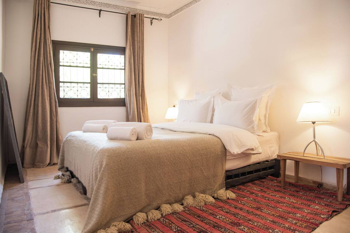 A neatly made bed with a patterned bedspread, a red and black striped rug, and a bedside lamp.