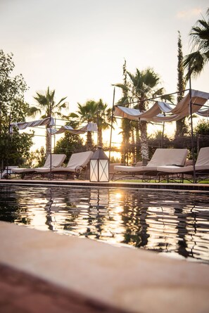 Un espace piscine agrémenté de chaises longues et de parasols, entouré de palmiers et avec un coucher de soleil en toile de fond.