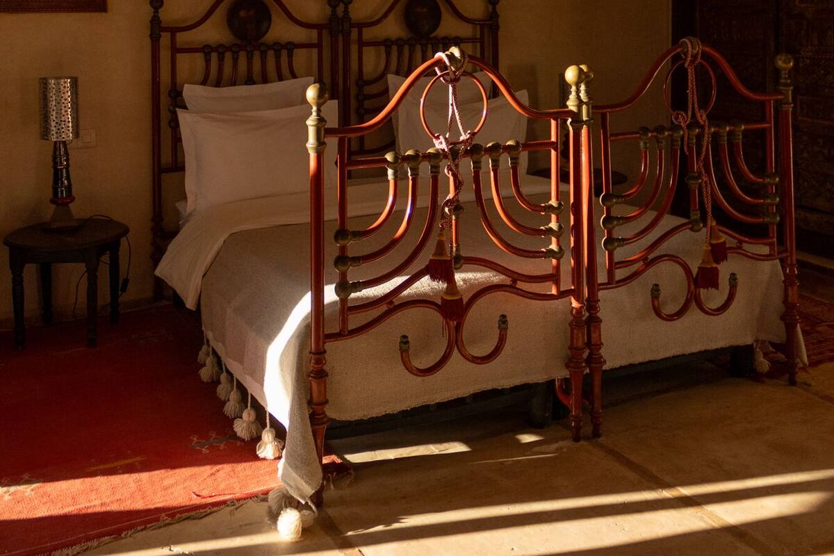 A traditional four-poster bed with a wooden frame and brass accents, positioned in a room with a red carpet and a small table with a lamp.