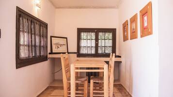 A small room with a wooden table and chairs, two windows with curtains, a framed picture on the wall, and a patterned rug on the floor.