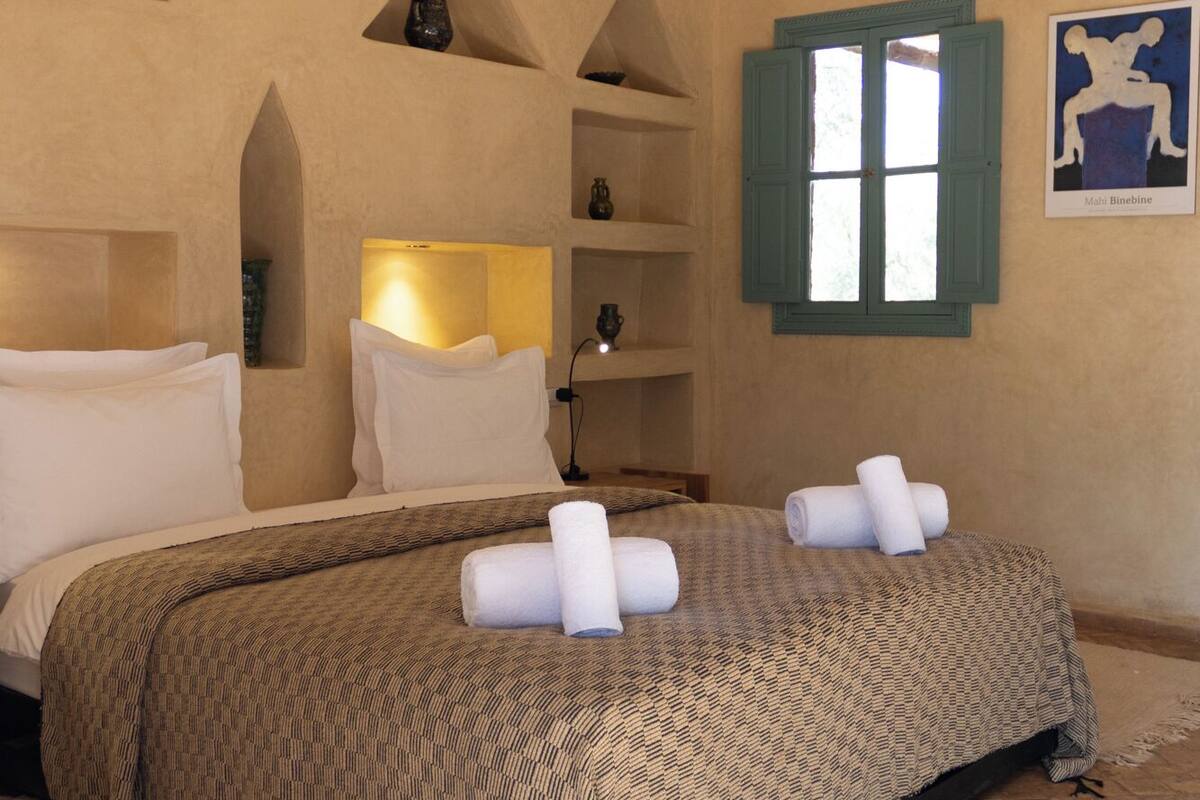 A bedroom with a bed, pillows, a nightstand, a shelf with decorative items, a window, and a painting on the wall.