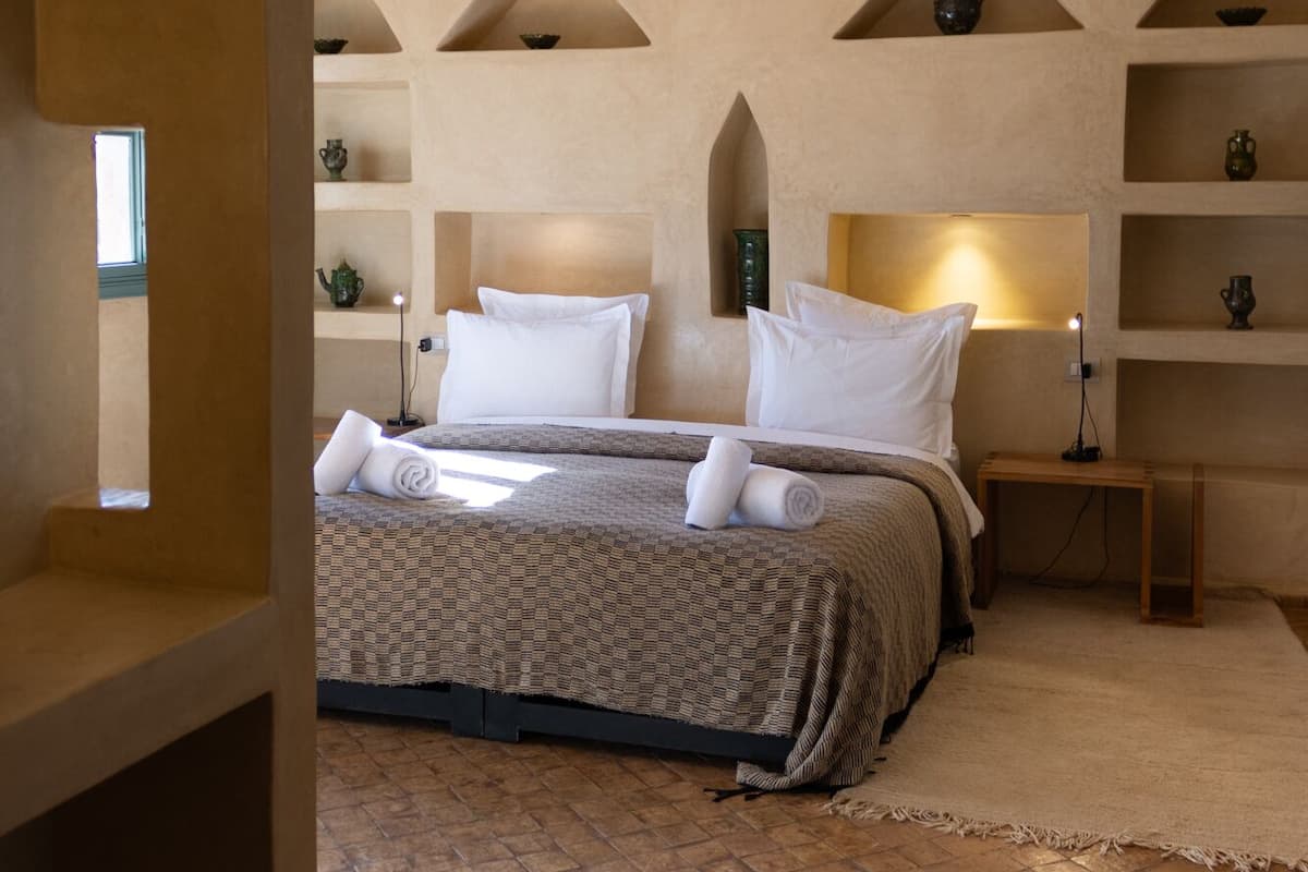 A bedroom with a high, vaulted ceiling, a large bed with a patterned bedspread, two bedside tables with lamps, and built-in shelves with decorative items.
